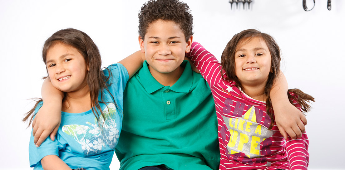 Three smiling children: two girls and a boy, with arms around each other. Girl on right wears SHINE LIKE A STAR shirt