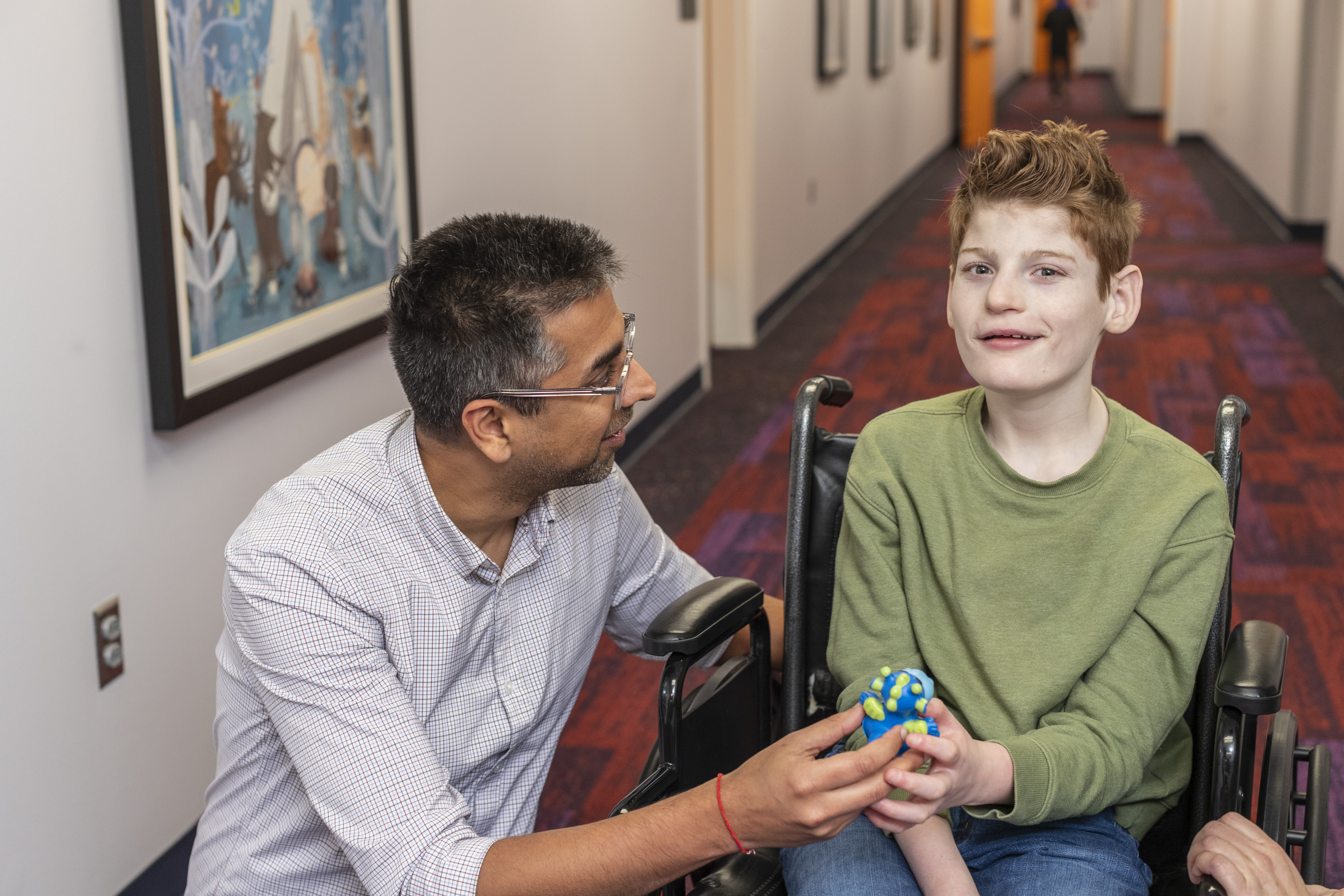 Young boy wearing a green shirt sitting in a wheelchair with doctor in glasses wearing a blue checkered shirt kneeling next to him